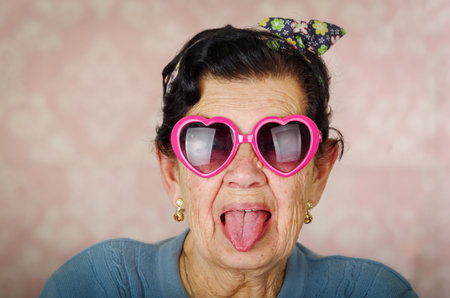 Older cool hispanic woman wearing blue sweater, flower pattern bow on head and pink heartshaped sunglasses looking into camera showing her tongue.の写真素材