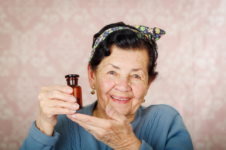 Older cool hispanic woman wearing blue sweater, flower pattern bow on head holding up a small red glass bottle and smiling to camera.の写真素材