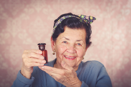 Older cool hispanic woman wearing blue sweater, flower pattern bow on head holding up a small red glass bottle and smiling to camera.の写真素材
