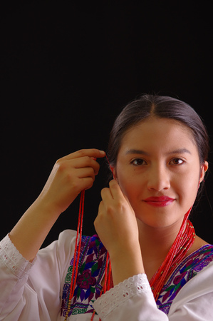 Beautiful young hispanic woman wearing white blouse with traditional embroided edges, attaching red hair extensions herself and smiling, black studio background.の写真素材