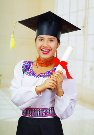 Young female student wearing traditional blouse and graduation hat, holding rolled up diploma, smiling proudly for camera.の写真素材