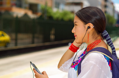 Pretty young woman wearing traditional andean blouse and blue backpack, waiting for bus at outdoors station platform, using mobile phone listening to music with headphones connected, smiling happily.の写真素材