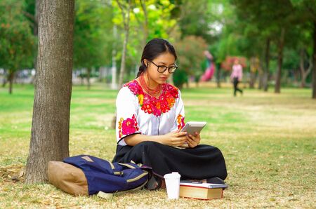Young woman wearing glasses, traditional andean skirt and blouse with matching red necklace, sitting on grass next to tree in park area, relaxing while using tablet, smiling happily.の写真素材