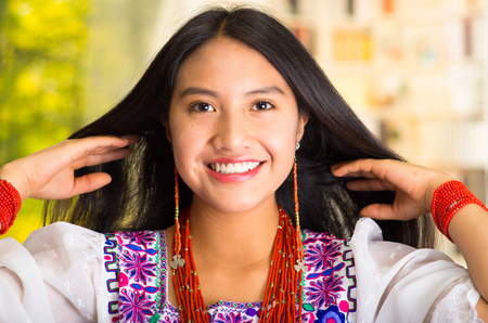 Beautiful hispanic woman wearing white blouse with colorful embroidery, lifting hair behind shoulders, smiling happily, garden background.の写真素材