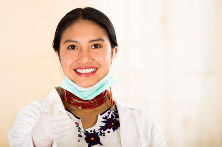 Young beautiful woman dressed in doctors coat and red necklace, facial mask pulled down to chin, smiling happily, egg white clinic background.の写真素材