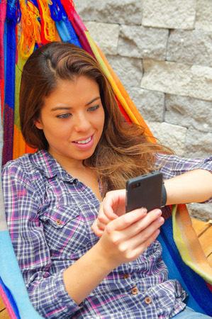 Woman wearing casual clothes holding mobile phone lying in hammock smiling, raising arm looking at smart watch, grey brick wall background.の写真素材
