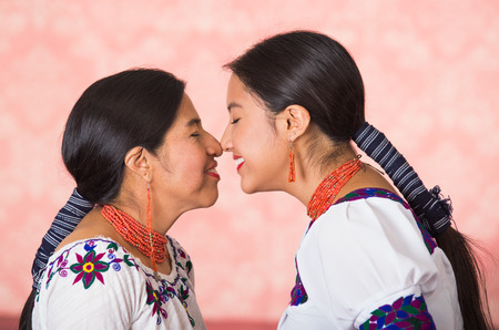 Beautiful hispanic mother and daughter wearing traditional andean clothing, seen from profile angle facing each other touching noses smiling, pink studio background.の写真素材