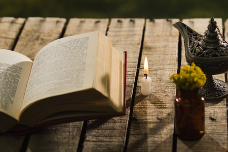Thick book lying open on wooden surface next to small brown bottle with yellow flowers and Aladin style lamp, beautiful night light setting, magic concept shoot.の写真素材