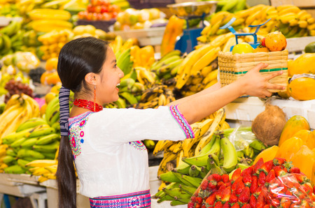 Beautiful young hispanic woman wearing andean traditional blouse posing for camera holding basket of fuits inside fruit market, colorful healthy food selection in background.の写真素材