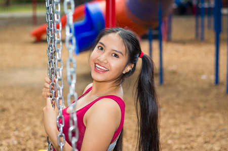 Young pretty teenage girl with pig tails wearing jeans and purple top, sitting on swing at outdoors playground, smiling happily for camera.の写真素材