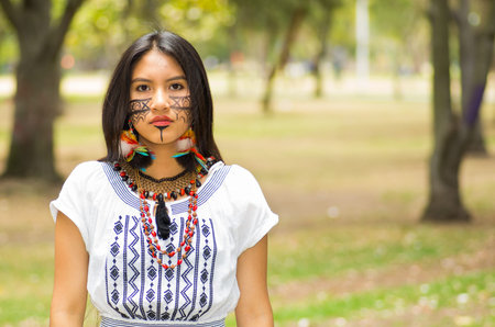 Beautiful Amazonian woman with indigenous facial paint and white traditional dress posing serious...