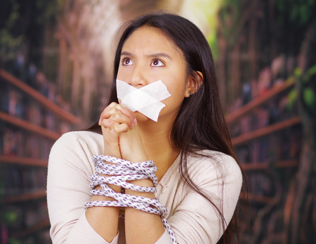 Young brunette woman tied up with rope around wrists and white tape covering mouth, facing camera, hostage concept.の写真素材