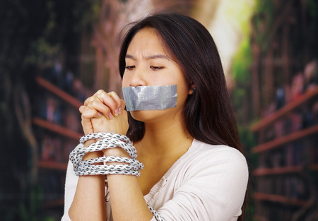 Young brunette woman wearing white sweater tied up with rope around wrists and duct tape covering mouth, facing camera, hostage concept.の写真素材