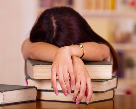 Young brunette woman lying bent over stack of books, appears to be asleep, tired student concept.の写真素材