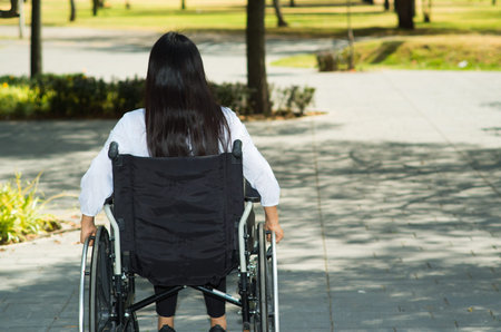 Young brunette woman sitting in wheelchair smiling with positive attitude, outdoors environment,...