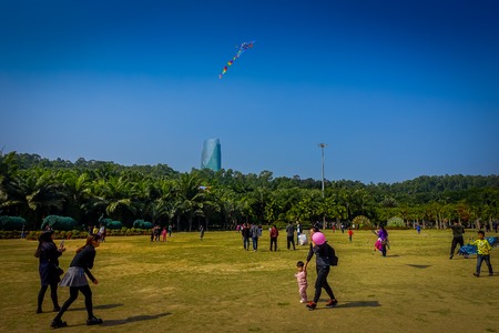 SHENZEN, CHINA - 29 JANUARY, 2017: Inside Lian Hua Shan park, large recreational area, people running on grass playing with kite, beautiful blue sky and tall building in background above treesのeditorial素材