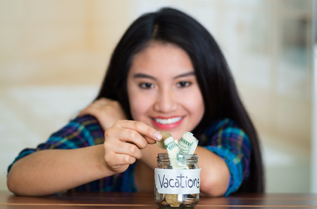 Young brunette woman sitting down facing camera, putting coins inside glass jar with money inside, label reading vacations, smiling happilyの写真素材