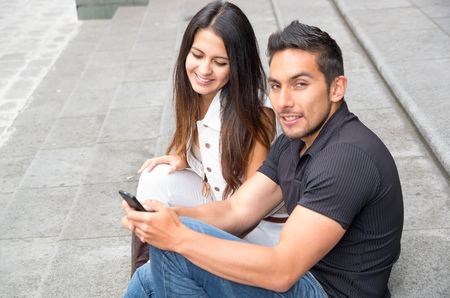 Charming young couple sitting on building steps using mobile phone and interacting happily, urban tourist conceptの写真素材