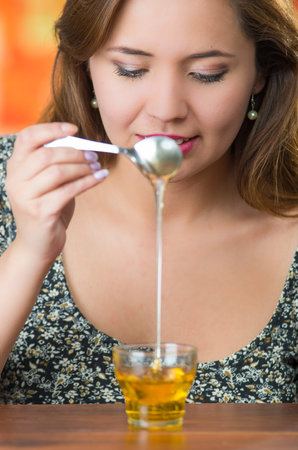 Young woman facing camera holding spoon with honey dripping into small glass on tableの写真素材