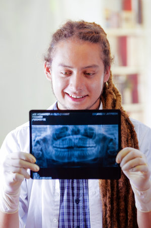 Young doctor with long dread locks posing for camera, holding up x ray image staring at it, clinic...