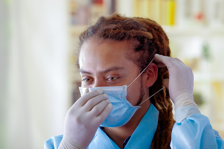 Young doctor with long dread locks posing for camera, adjusting facial mask covering mouth, clinic in background, medical conceptの写真素材
