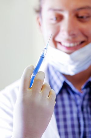 Young doctor with long dread locks posing for camera holding syringe, wearing facial mask covering mouth, clinic in background, medical conceptの写真素材