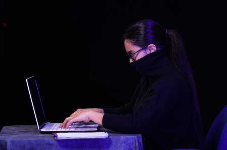 Portrait of a beautiful and smart girl hacker with laptop on dark background using glasses and covering her neck, mouth and nose with her turtleneck sweater while she is workingの写真素材