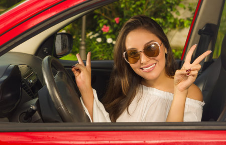 Pretty girl wearing sunglasses and doing victory signal with her both hands inside of her red car