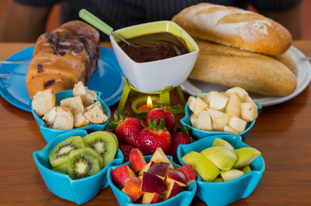 Delicious chocolate fondue inside of a white bowl with assorted fruits as, strawberry, pear, kiwi, apple and pieces of bread in bowls on wooden tableの写真素材