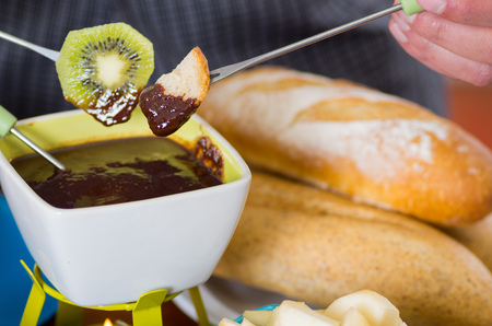 Delicious kiwi and a piece of bread in a metal stick covered with chocolate fondue inside of a white bowl on wooden tableの写真素材