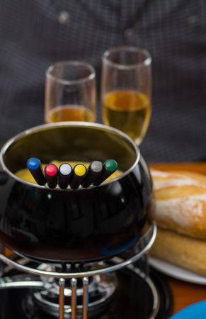 Gourmet Swiss fondue dinner with assorted cheeses and a heated pot of cheese fondue with colorful forks dipping inside of the pot, with white wines cups behind on a wooden tableの写真素材