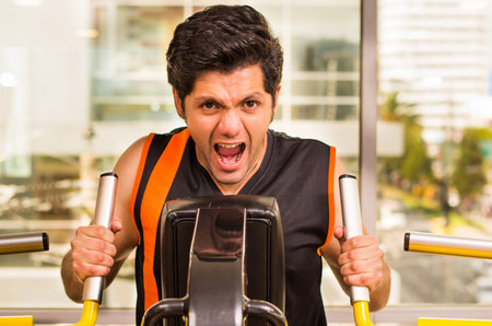 Handsome young man working on fitness machine at gym and screaming at same time