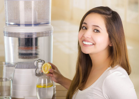 Beautiful smiling woman holding a cut lemon in her hand with a filter system of water purifier on a...