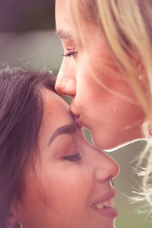 Close up of a lovely lesbian couple, blonde woman kissing in a forehead of a brunette girl, in a blurred backgroundの写真素材