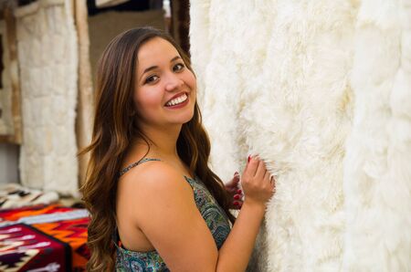 Close up of a beautiful smiling young woman touching a white soft andean traditional blanket clothing and posing for camera, colorful fabrics backgroundの写真素材