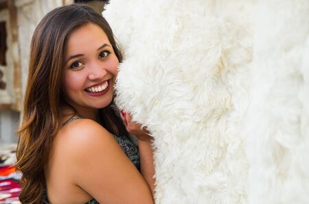 Close up of a beautiful smiling young woman touching a white soft andean traditional blanket clothing and posing for camera, colorful fabrics backgroundの写真素材