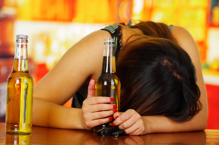 Portrait of a drunk woman sleeping over a wooden table and holding an open beer with her hand, in bar backgroundの写真素材