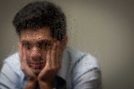 Worry depresed young man with both hands in his face, behind a blurred window with drops, gray backgroundの写真素材