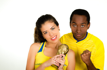 QUITO, ECUADOR - MAY 06, 2017: Young ecuadorian couple wearing official Marathon football shirt standing facing camera, and girl is holding a world cup trophy in a white backgroundのeditorial素材