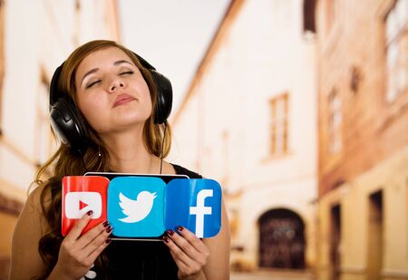 Quito, Ecuador - March 11, 2016: Woman holding a printed stickers of facebook, youtube and twitter icons with both hands and wearing a black headphones, in blurred city backgroundのeditorial素材