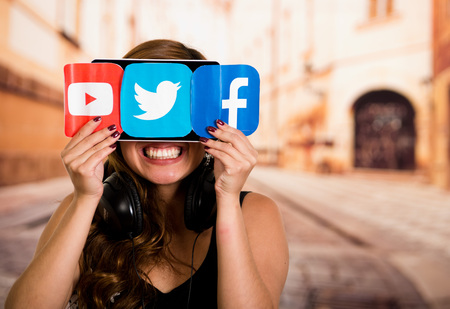 Quito, Ecuador - March 11, 2016: Smiling woman hiding behind a printed stickers of facebook, youtube and twitter icons with both hands in blurred city backgroundのeditorial素材