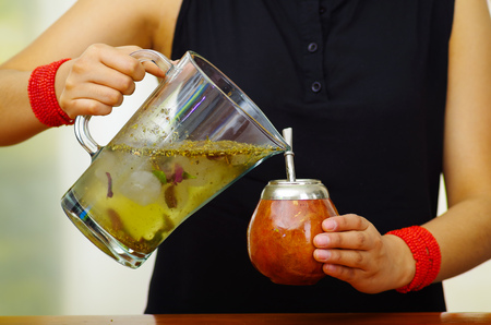 Woman holding trasnparent jar containing water and green herbs, pouring into traditional cup with typical metal straw sticking up, preparing popular drink called mateの写真素材