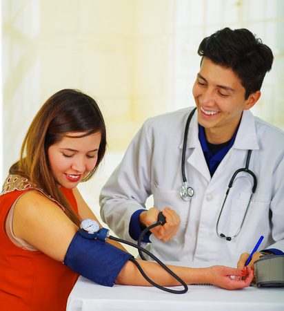 Close up of a handsome smiling young doctor with stethoscope around his neck taking the pulse to a young woman with a tensiometer, in a doctor consulting room backgroundの写真素材