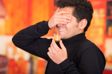 QUITO, ECUADOR- 17 OCTOBER, 2015: Close up of a smiling young man holding in his hand a golden gift present and covering his eyes with his hand, wearing a black turtleneck jacket in a blurred background.のeditorial素材