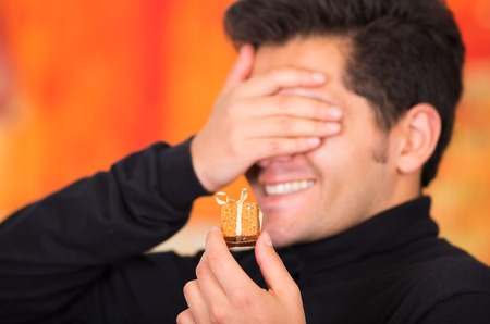 QUITO, ECUADOR- 17 OCTOBER, 2015: Close up of a smiling young man holding in his hand a golden gift present and covering his eyes with his hand, wearing a black turtleneck jacket in a blurred background.のeditorial素材