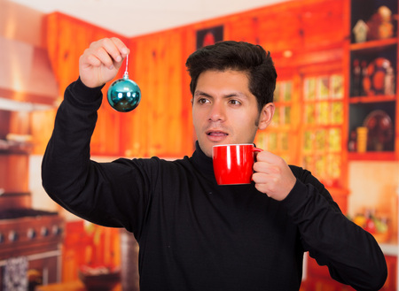 QUITO, ECUADOR- 17 OCTOBER, 2015: Close up of a handsome young man holding in his hands a beautiful blue ball christmas tree decoration with one hand and with his other hand a red cup in a blurred background.のeditorial素材