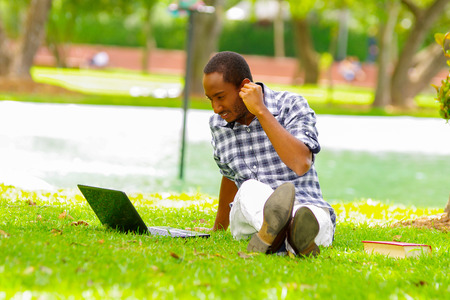 Young black tourist man sitting down on green grass, listening music with his headphones and checking his computer in the city of Quito Ecuadorの写真素材