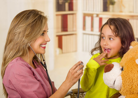Young pretty girl smiling and hugging her teddy bear while a beautiful smiling doctor examining with a stethoscope in a office background.の写真素材