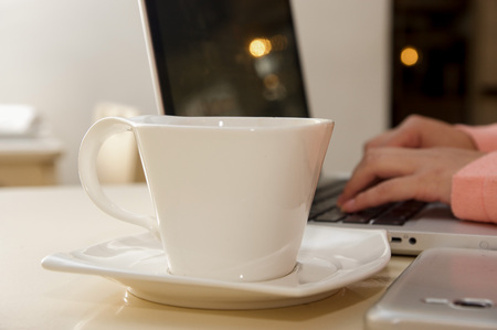Close up of a white cup of coffe with a blurred businesswoman hand working in the background. Business conceptの写真素材