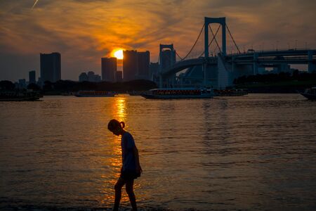 TOKYO, JAPAN JUNE 28 - 2017: Unidentified woman with a shadow walking in Odaiba Beach at Sunset, with the Osaiba bridge in the horizont, Tokyoのeditorial素材